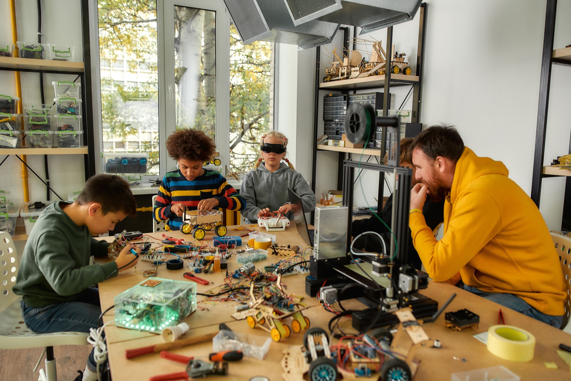 Learning, friendship and fun. Young technicians building robots and vehicles, using soldering iron to join chips and wires, testing toys together with a male teacher at a stem robotics class